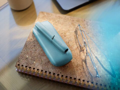 Turquoise IQOS Originals Duo device on a notebook next to glasses.