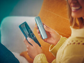 Hands holding a mobile phone and IQOS Originals One device, downloading the app to get the most of the heated tobacco device.